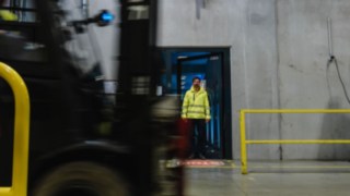 Muž v ochranném oděvu stojí ve dveřích tovární haly. Okolo projíždí vysokozdvižný vozík. Muž v ochranném oděvu stojí ve dveřích tovární haly. Okolo projíždí vysokozdvižný vozík.