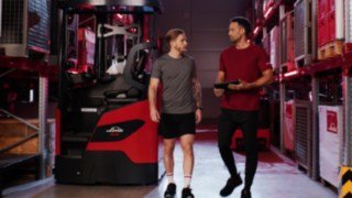 Dvě osoby ve sportovním oblečení stojí před vysokozdvižným vozíkem se zvedacím sloupem Ri16 od společnosti Linde Material Handling, jedna z osob drží tablet.